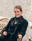 A smiling woman sits in a woven chair outdoors, wearing a black moto-style jacket, black pants, and a bright rainbow-striped neck gaiter. She has sunglasses on her head and a bicycle wheel visible on the right side of the frame.