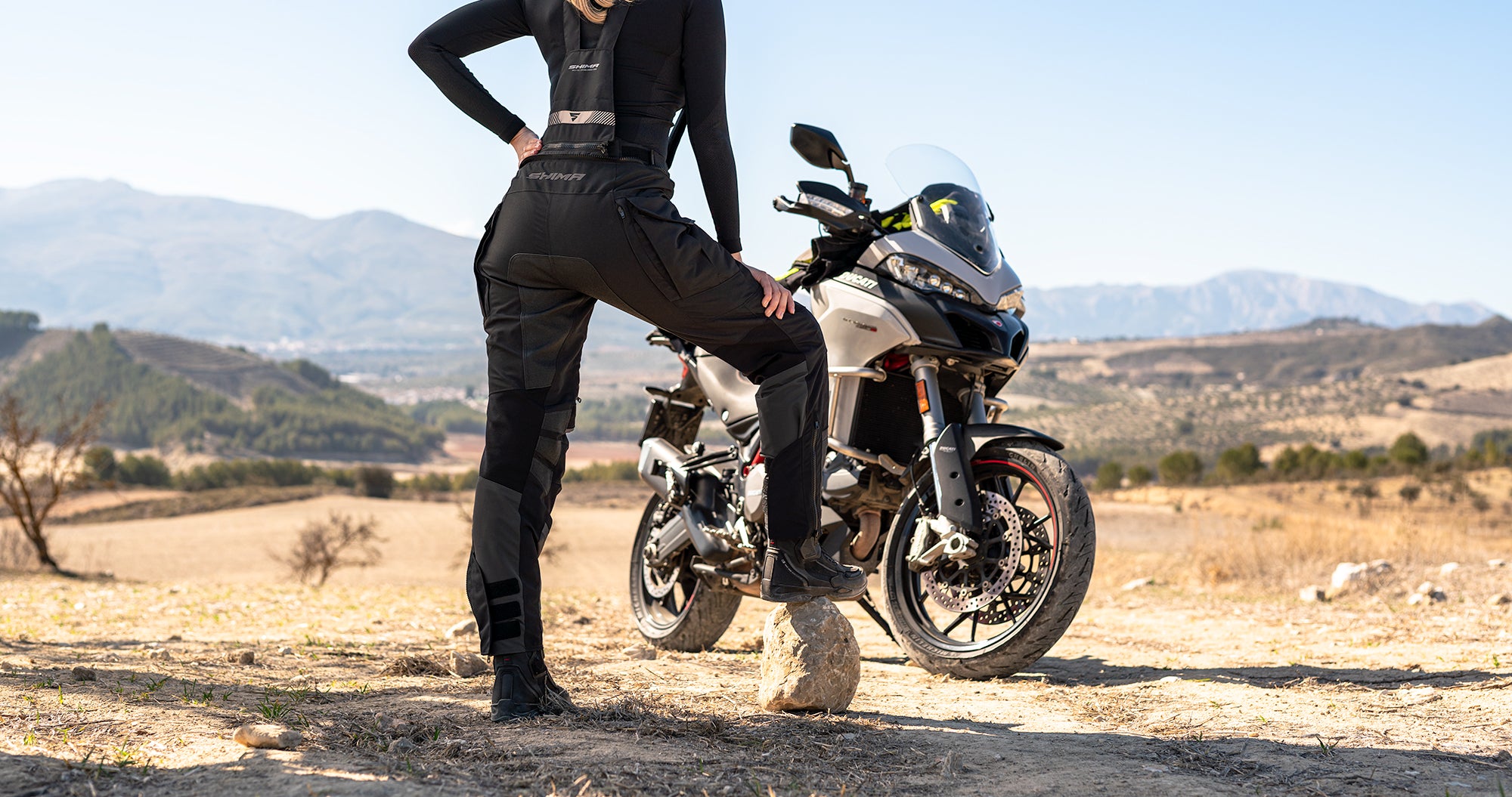 A woman standing by her motorcycle wearing Black SHIMA touring motorcycle pants for women 
