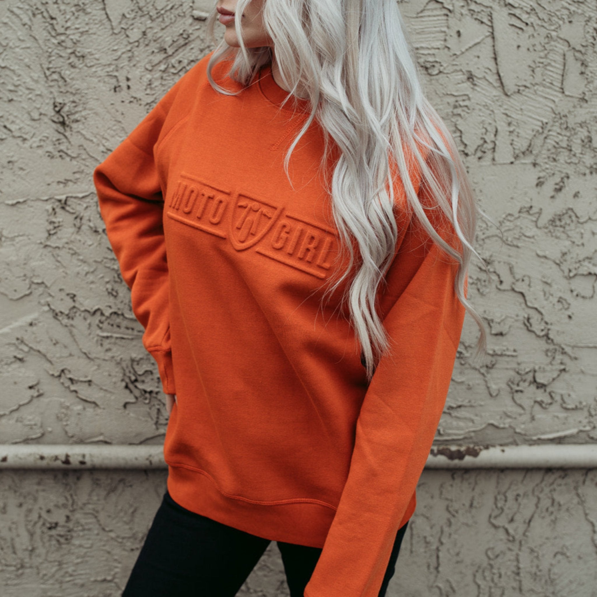 A young woman standing by a wall and wearing orange colour lady sweatshirt with Moto Girl 3D logo