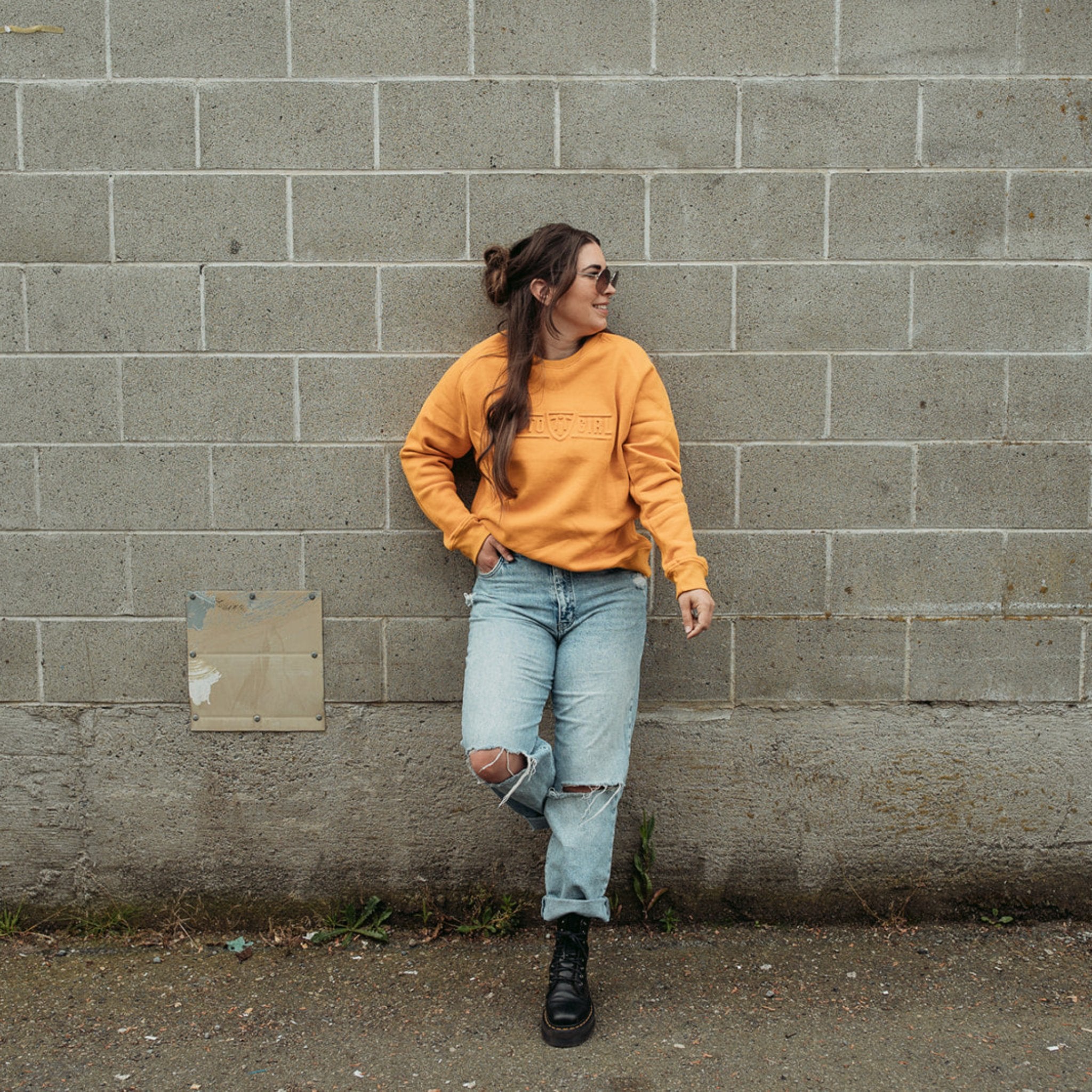 A young woman standing by a wall and wearing yellow/mustard colour lady sweatshirt with Moto Girl 3D logo