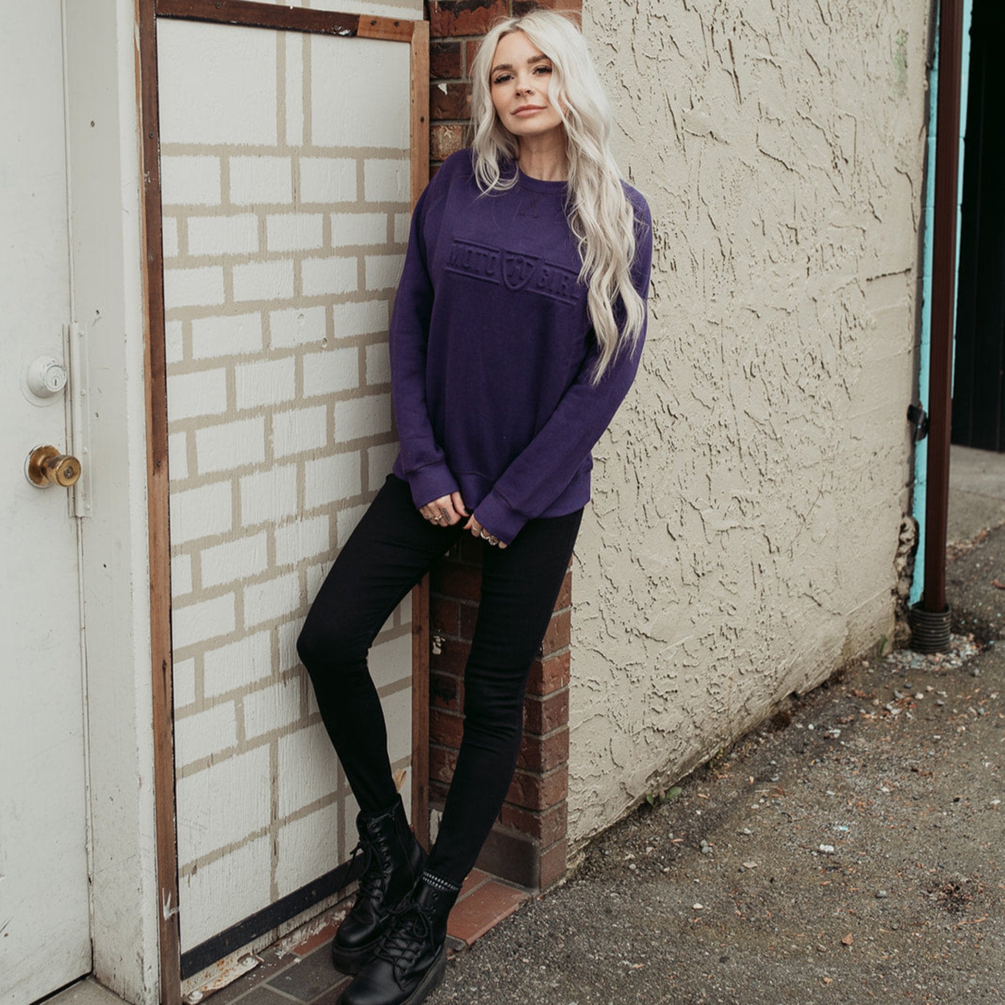 A young woman standing by a wall and wearing purple colour lady sweatshirt with Moto Girl 3D logo