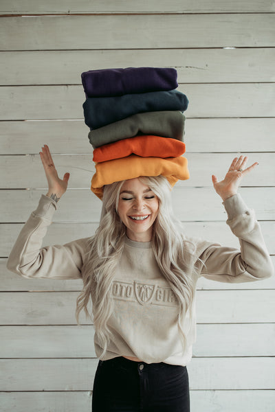 a young woman wearing Sand colour lady sweatshirt with Moto Girl 3D logo and holding 5 colourful sweatshirts on her head
