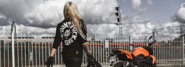 A blond woman walking towards the orange motorcycle wearing Pando Moto black sweatshirt with while "Don't Fucking Die" motive on the back