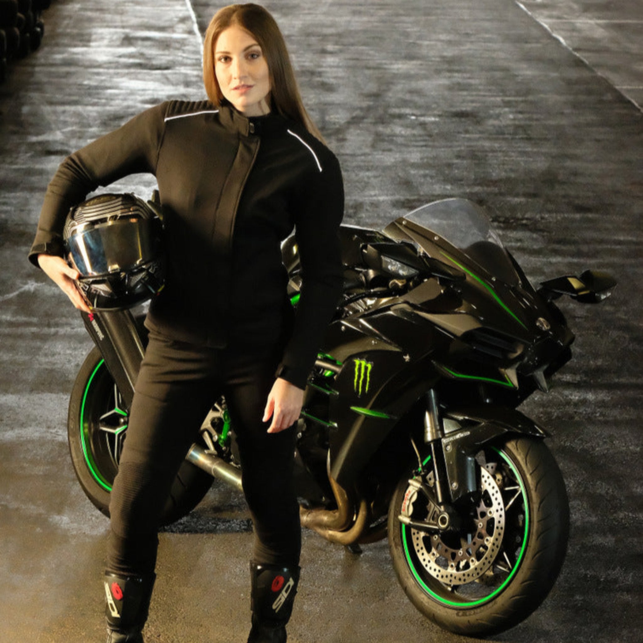 A young woman standing by the motorcycle wearing black motorcycle protective clothes