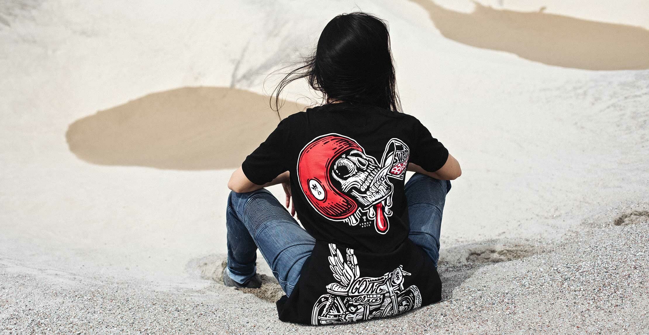 A woman siting on the beach wearing black Pando Moto women&#39;s motorcycle t-shirt with red scull motive