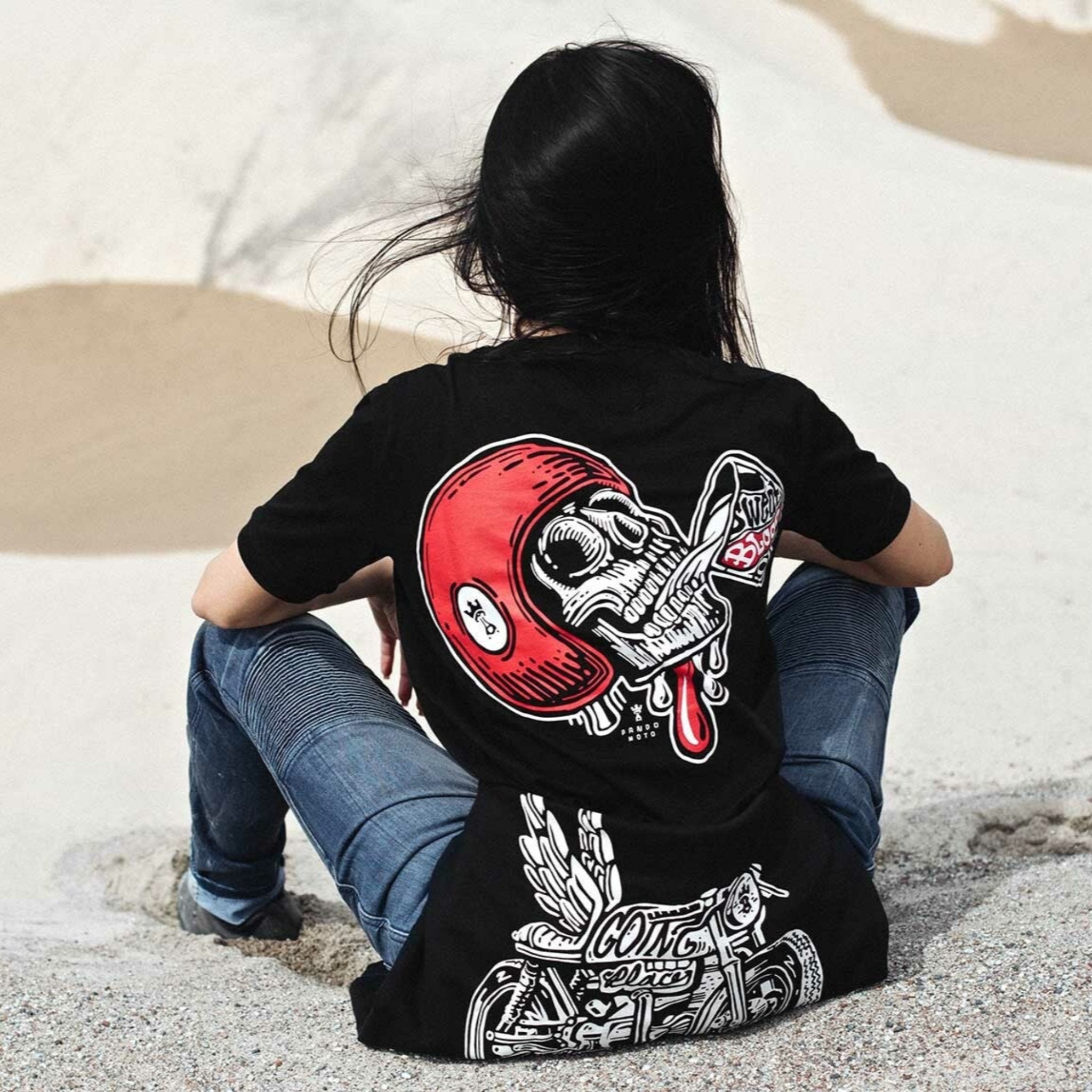 A woman siting on the beach wearing black Pando Moto women&#39;s motorcycle t-shirt with red scull motive