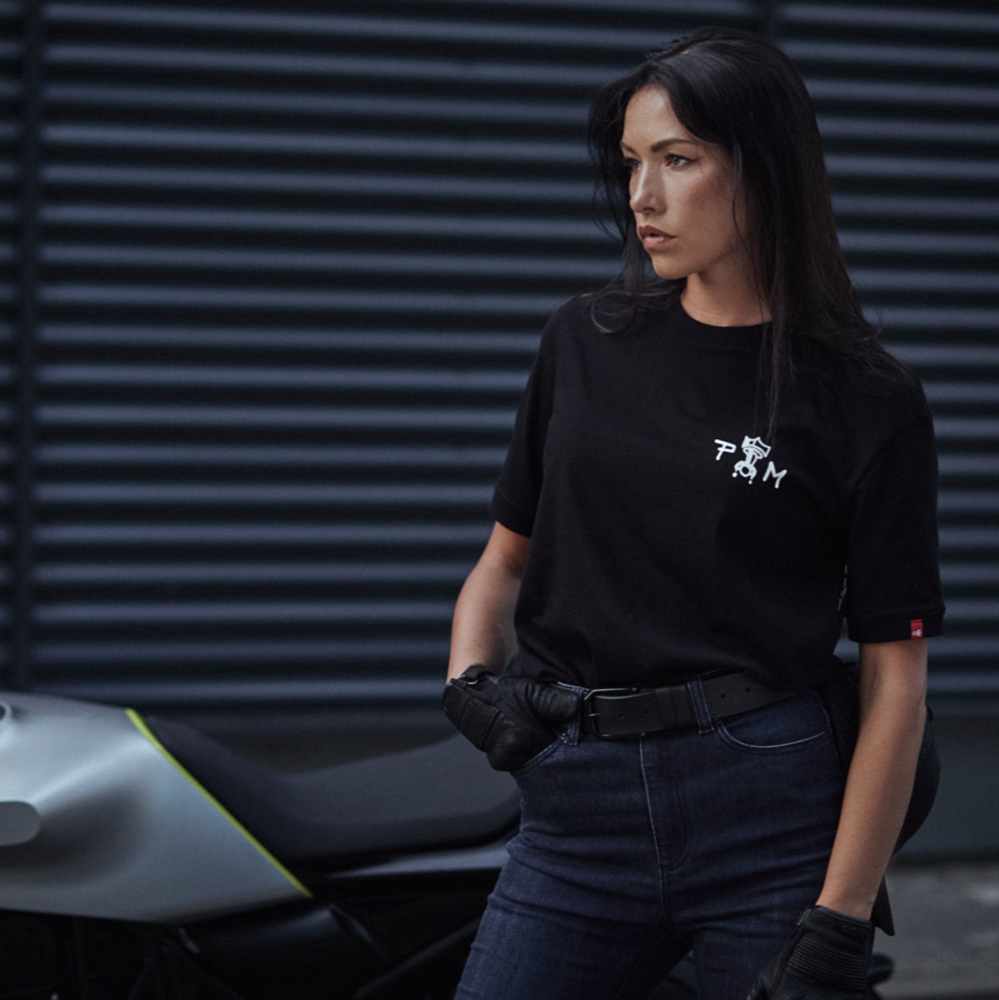 A young woman standing by her motorcycle wearing blue high waist motorcycle jeans and pando moto t-shirt