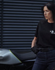 A young woman standing by her motorcycle wearing blue high waist motorcycle jeans and pando moto t-shirt