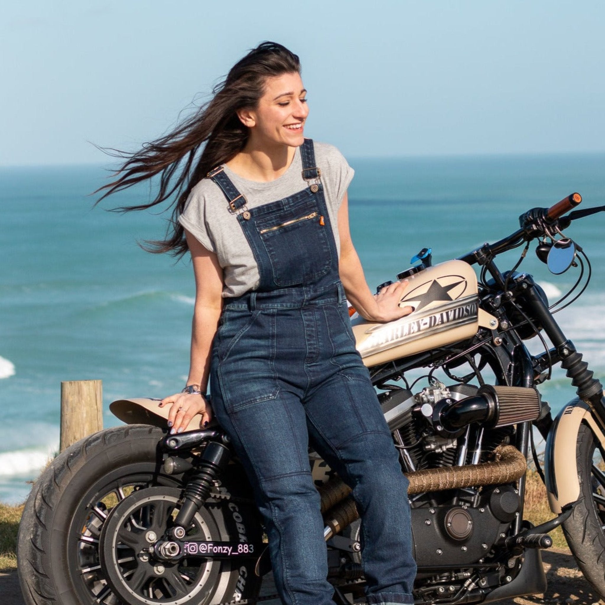 A woman by the beach with her motorcycle wearing blue women's motorcycle overall from Moto Girl with zipper pocket detail on the chest