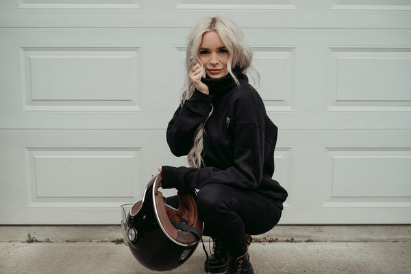 A young blond women wearing high neck black sweatshirt with motogirl logo in front