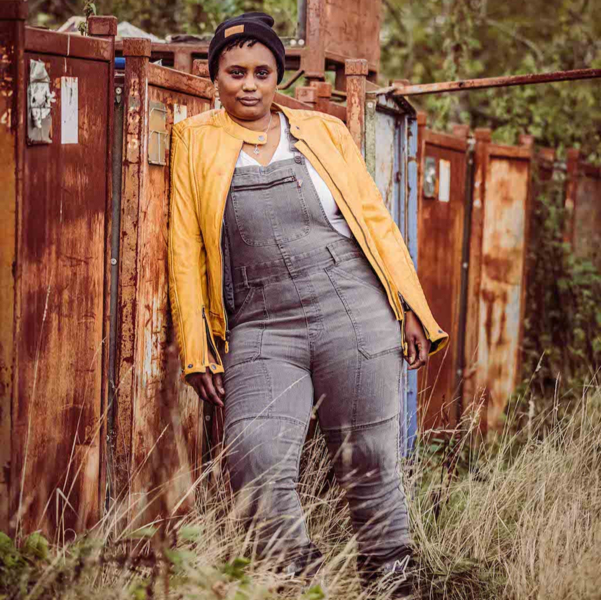 A woman wearing grey motorcycle overall and yellow leather jacket standing by the metal container