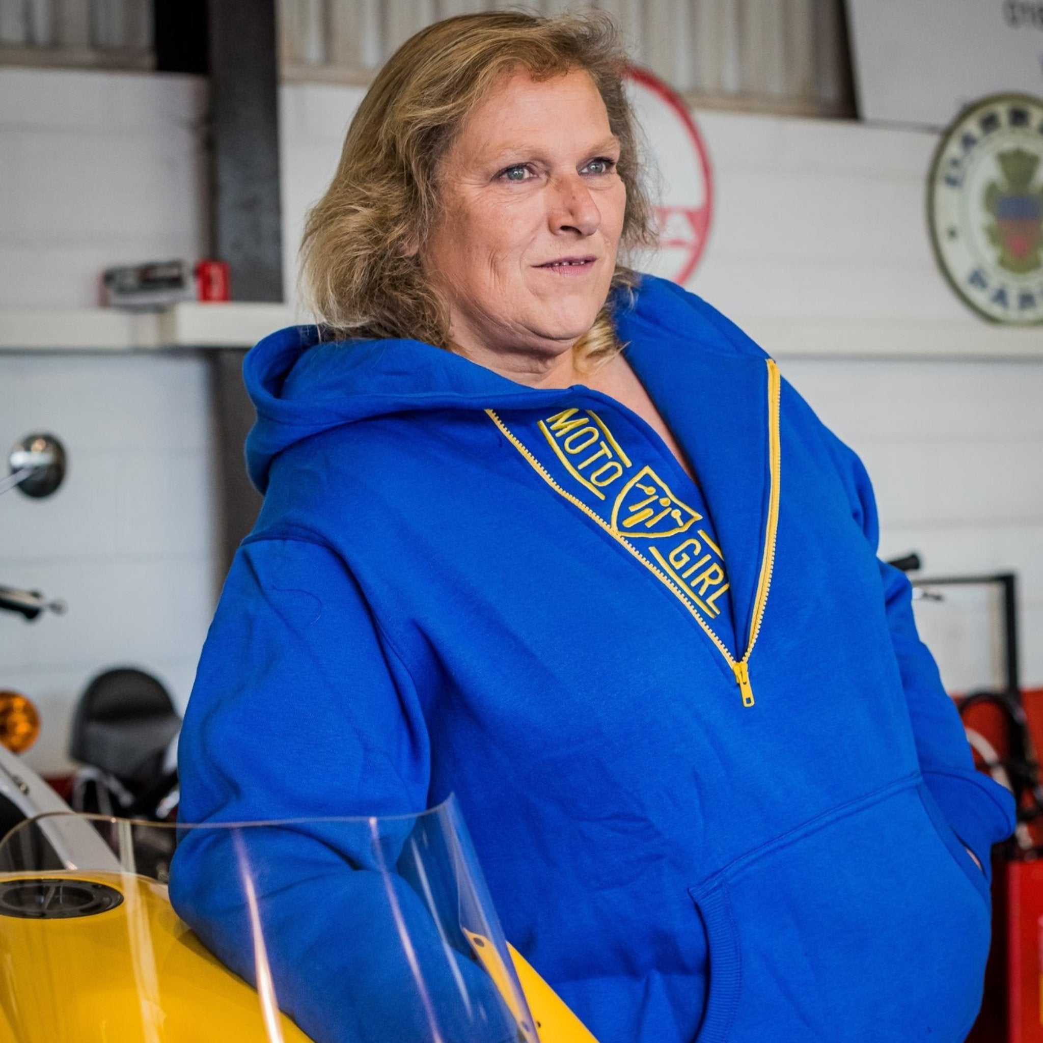 A blond woman standing by a motorcycle wearing blue and yellow motorcycle hoodie from Moto Girl