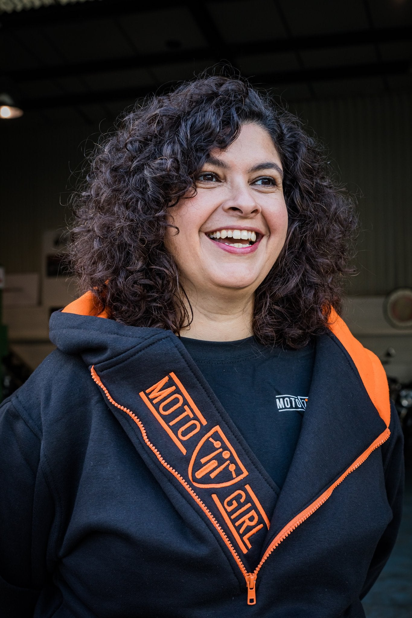 A smiling woman wearing Black and orange motorcycle hoodie from MotoGirl with a long zip