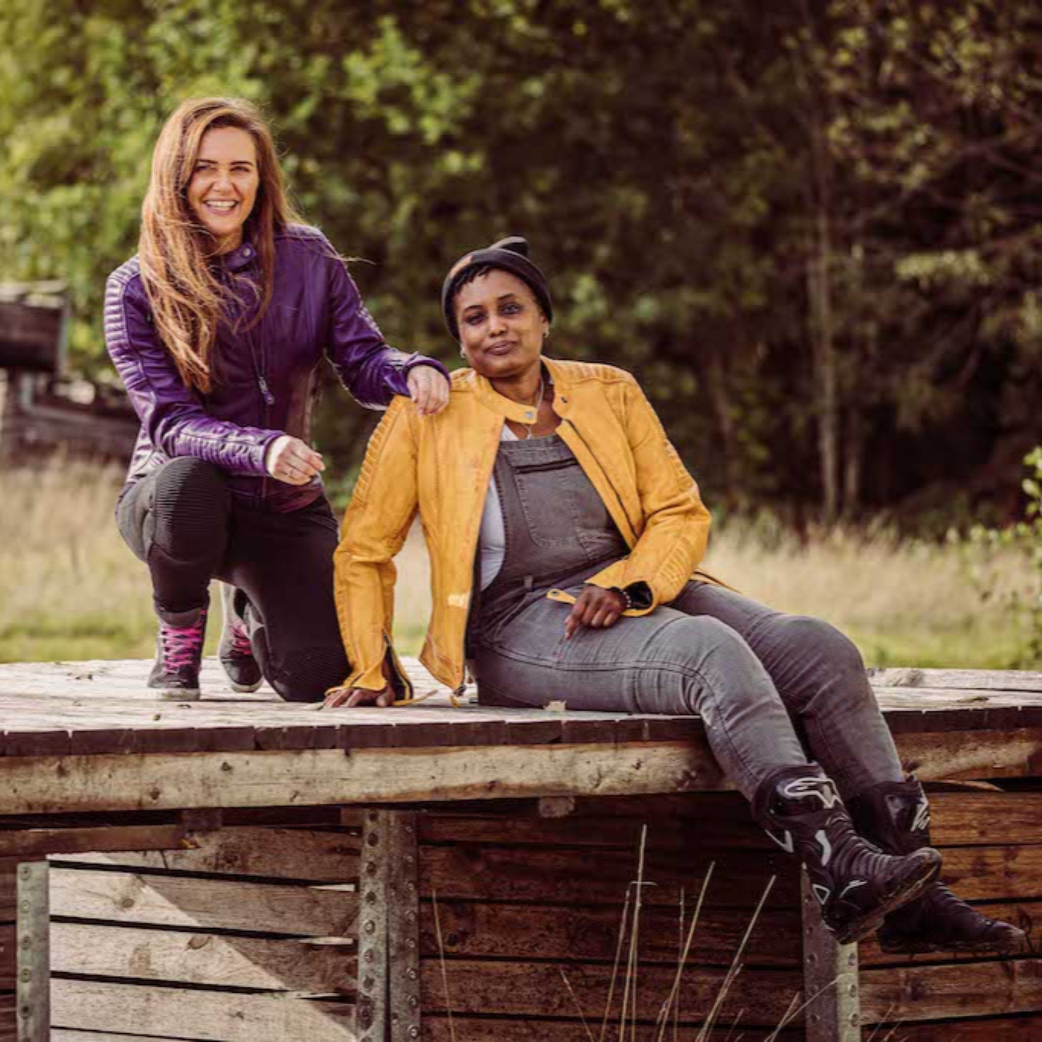 two smiling women sitting outside wearing motorcycle jackets in yellow and purple from moto girl