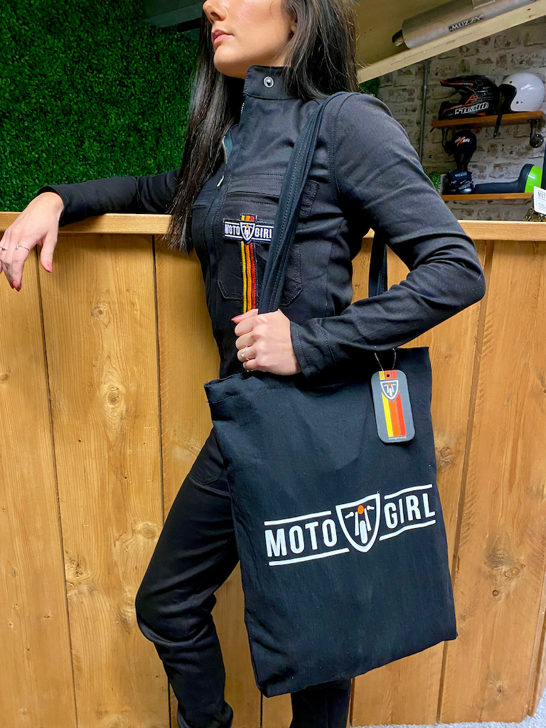 A woman leaning on the shop counter wearing black women&#39;s garage suit  from MotoGirl and a MotoGirl bag