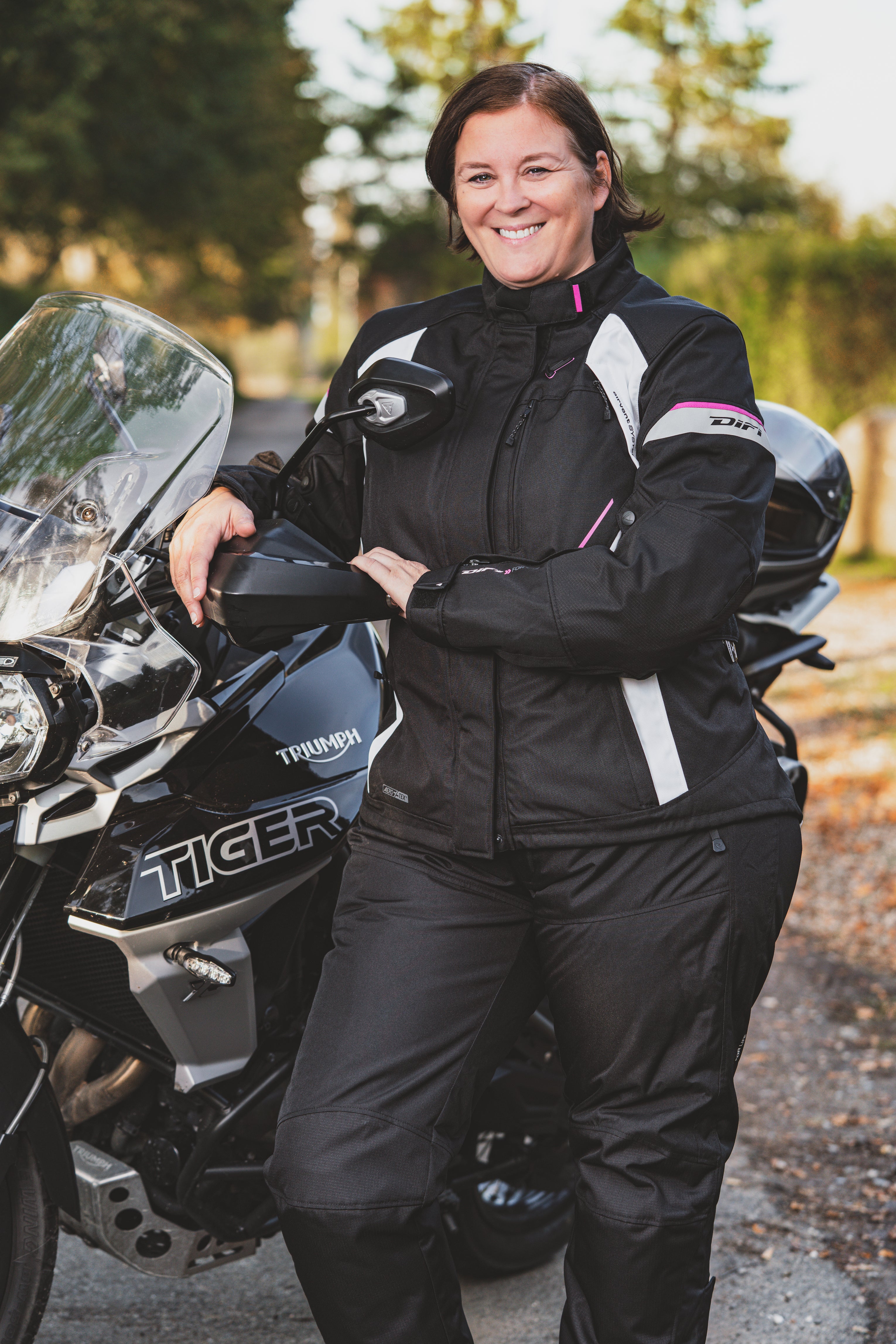A smiling woman standing by her Triumph Tiger motorcycle wearing Difi textile women's motorcycle Nexia jacket and Cyclone pants