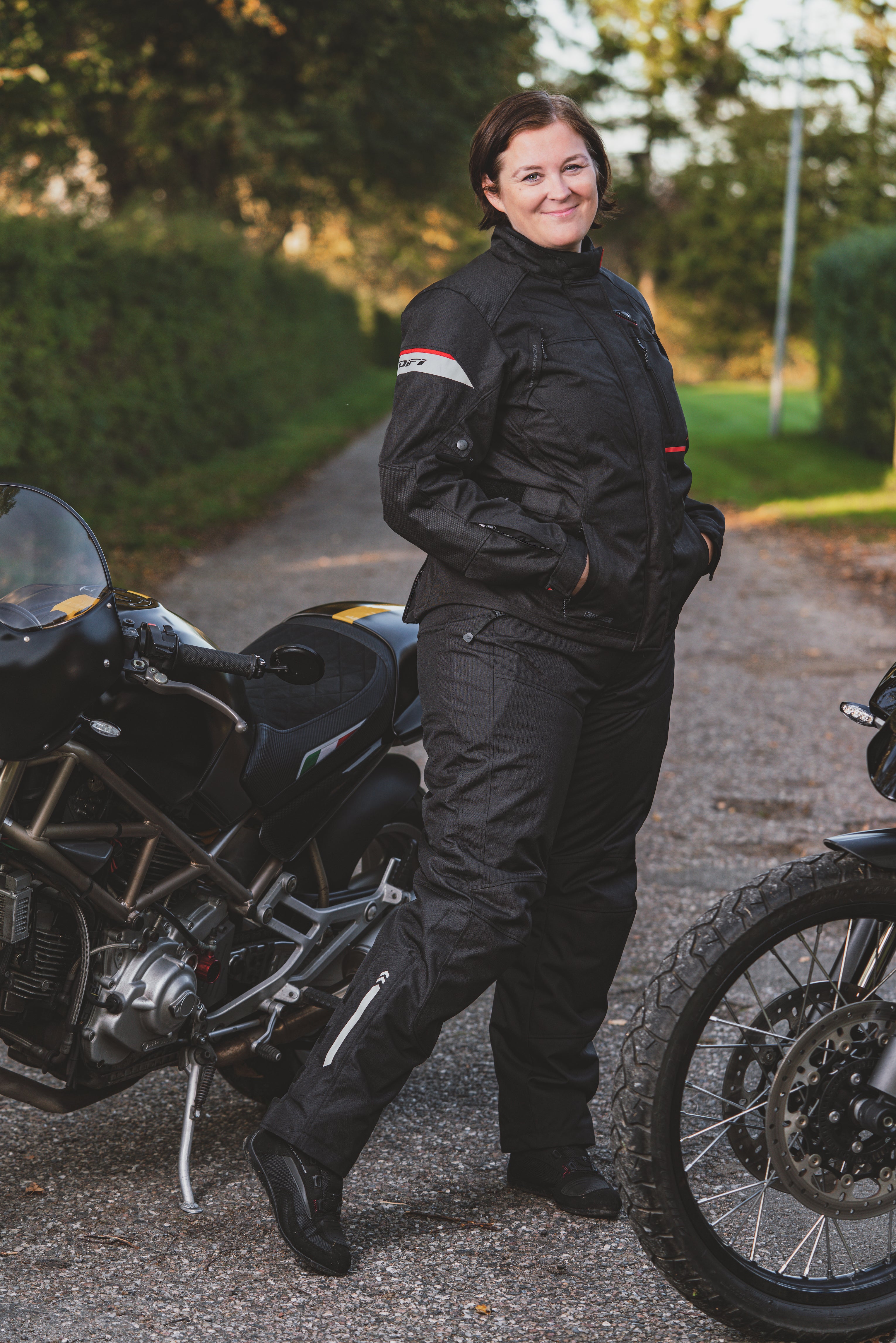 A smiling woman standing by motorcycles wearing Difi textile women's motorcycle Nexia jacket and Cyclone pants