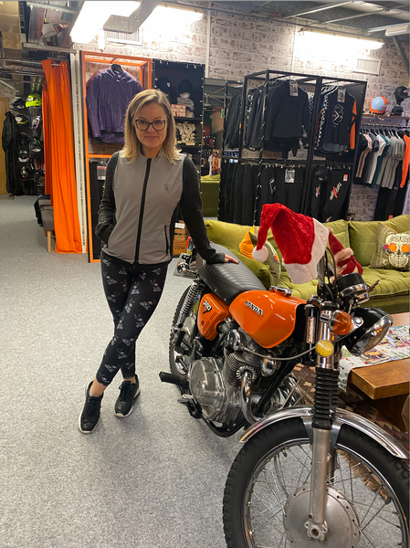 A blond woman leaning on the orange motorcycle wearing a grey vest